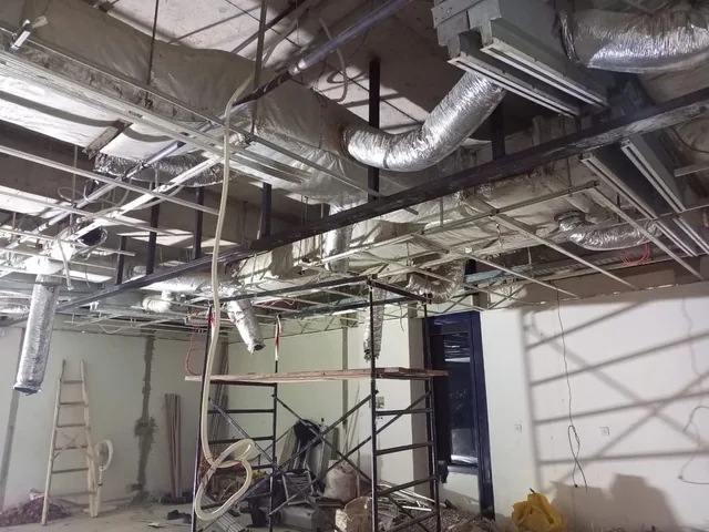 Wide room view looking up at exposed insulated ducts, metal ceiling framing and suspended wires above scaffolding.