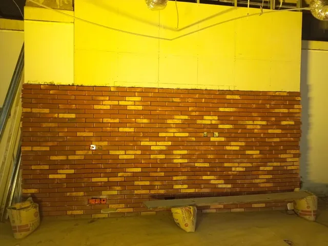 Red and pale brick-faced wall under yellow work lighting, with buckets and a low platform at its base.