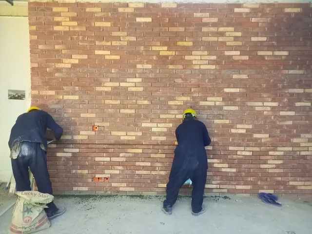 Two workers facing a broad wall of mixed red and pale bricks, with a material bag and tools on the floor.