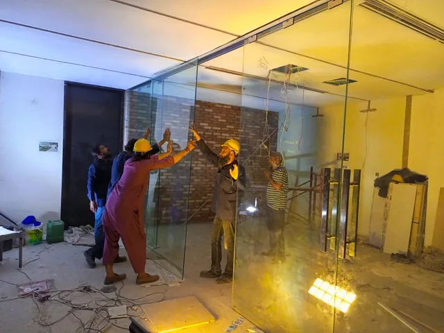 Workers beside full-height clear glass panels with a brick wall behind, floor cables and bright temporary work lights.