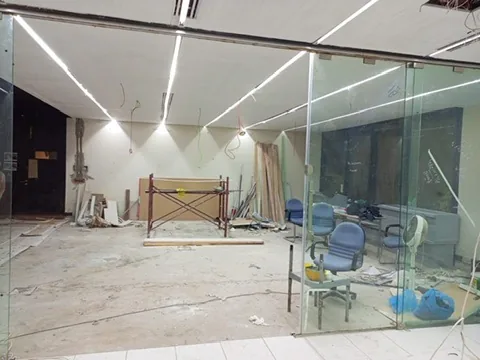 Room with bright linear ceiling lights, glass partitions, a small scaffold, loose materials and chairs.