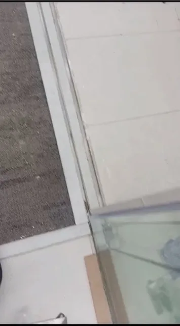 Close downward view of a glass partition edge and metal floor track beside pale tile and gray flooring.
