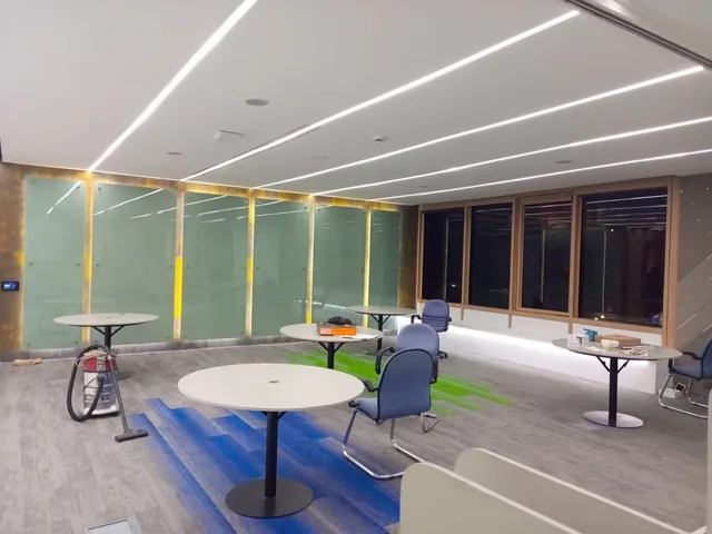 View into a room with round tables, linear ceiling lights, illuminated vertical wall strips, dark windows and a vacuum cleaner.