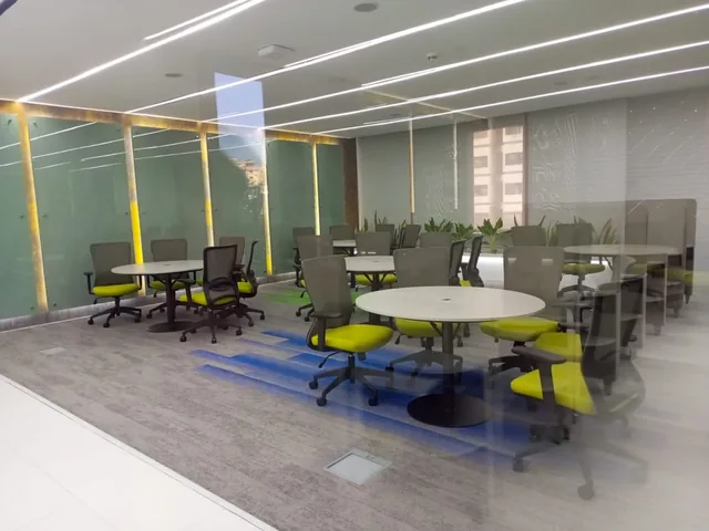 Glazed room containing round tables with lime-green and gray chairs, with a line of individual seating enclosures at the rear.