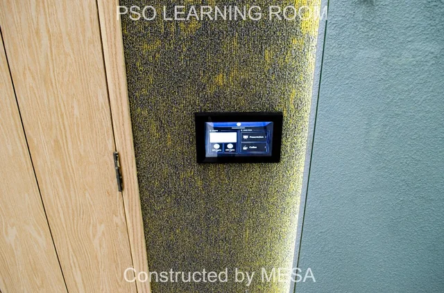 Small black-framed touchscreen on a textured gold-gray wall panel beside a timber-toned door and gray wall, retaining original overlays.