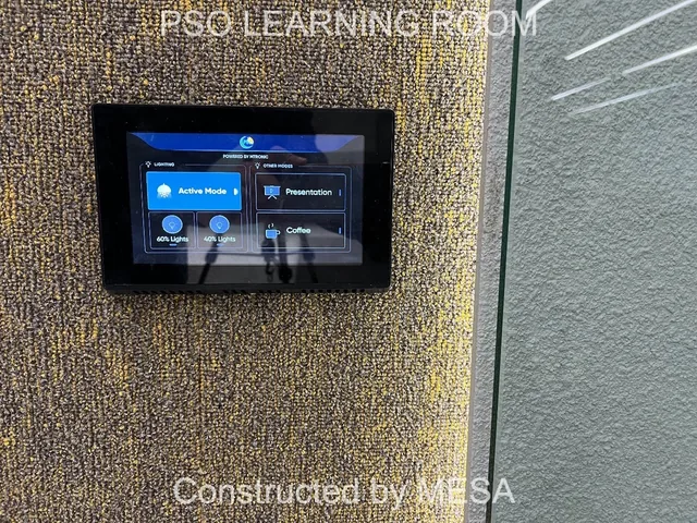 Close view of a black-framed touchscreen with a blue interface on a textured wall, retaining original PSO Learning Room and MESA overlays.