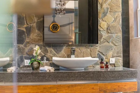 White vessel basin on a dark counter above timber-toned drawers, with a stone backdrop and mirror reflecting the bedroom.