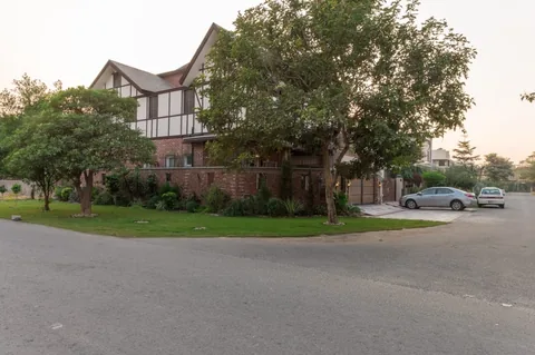 Wide street-corner view of the house with pale gables, dark facade lines, brick boundary walls, mature trees and lawn.