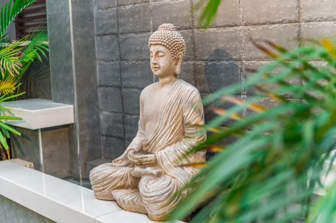 Pale seated Buddha sculpture on a ledge against gray masonry, framed by green palm leaves.