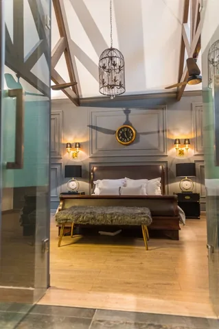 Full-height bedroom view with a pitched white ceiling, dark exposed roof members, hanging cage pendant, panelled wall and centred bed.