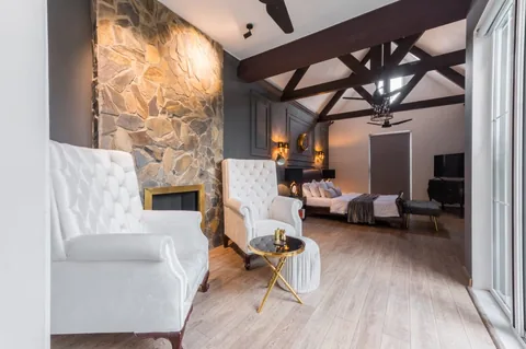 Long bedroom with white wing chairs and a stone fireplace in the foreground, a bed beyond and dark roof members overhead.
