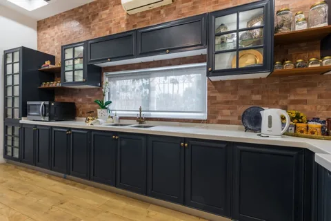 Kitchen with deep blue framed cabinets, brass-coloured knobs, white countertop, brick wall, display cupboards and a window above the sink.