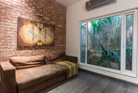 Small sitting room with brown sofa, brick wall and map artwork beside a large window onto a narrow planted stone court.