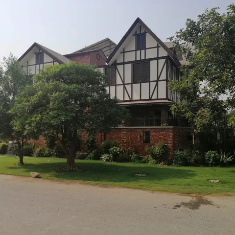 Daytime exterior with two pale gables, dark facade lines, brick base and boundary, mature garden trees and visible rooftop equipment.