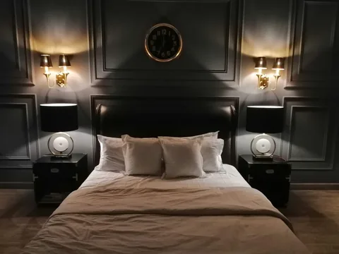 Bedroom at night with warm paired sconces and bedside lamps illuminating gray wall panelling, a dark headboard and pale bedding.