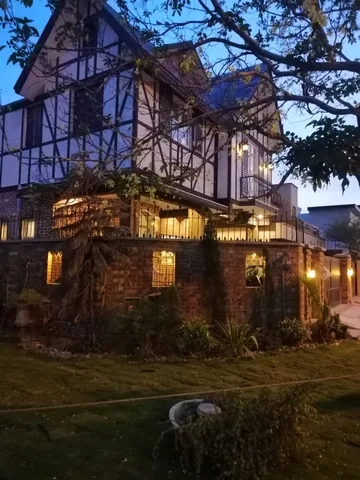 Portrait exterior at dusk with pale gables, dark facade lines, warm porch and boundary lanterns, trees and blue sky.