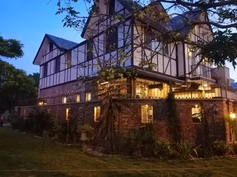Wide dusk view of the gabled house behind trees and a brick boundary, with warm lanterns and interior light against the blue evening sky.
