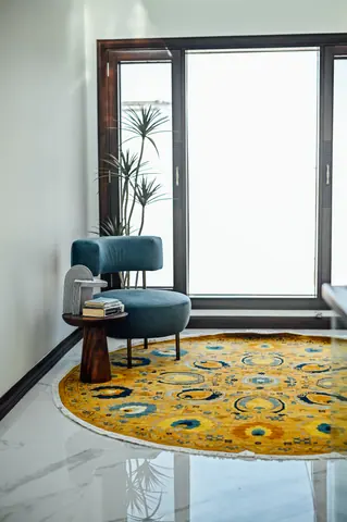 Blue armchair, timber side table and plant on a yellow circular rug beside a tall window.