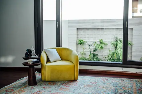 Yellow armchair and timber side table beside a large window overlooking a planted boundary wall.