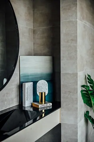 Curved mirror above a black bathroom counter holding small sculptures, books and artwork beside grey stone tiles.