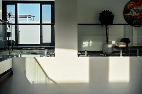 Sunlight across a white atrium wall below an upper gallery with a window, tree and bench.