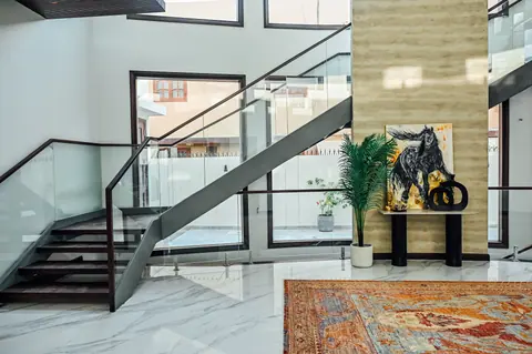 Wide view of an open dark stair crossing a bright hall beside a pale stone wall, plants and artwork.