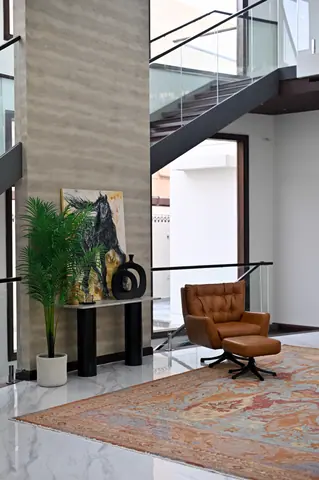 Brown lounge chair and footstool beneath an open stair beside a tall stone wall and console.