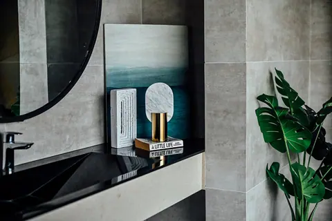 Black vanity counter, round-edged mirror, grey stone wall and decorative objects with a leafy plant.